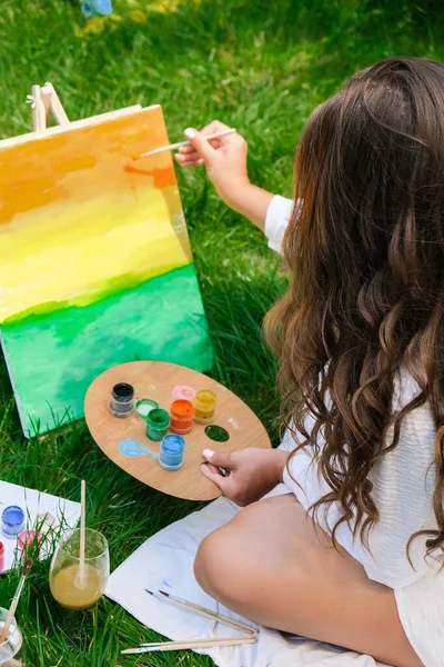 Artist painting on the easel outdoors in the garden. Open air outdoor art workshop. Draw on the canvas with brush and palette sitting on the grass during a picnic in a park. Creativity, art and hobby concept