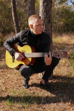 Gülümseyen beyaz ressamın portresi sonbahar parkında ahşap akustik gitar çalar. Genç söz yazarı, sonbahar zamanı dışarıda telli müzik aleti hobisi yapıyor. Sesli müzik doğal olarak iyileşir.