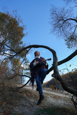 Gülümseyen beyaz kızın portresi sonbahar parkında ukulele çalıyor. Genç kadın, sonbahar zamanı dışarıda gitar enstrümanı çalıyor. Ses müziği doğal çevrede iyileşiyor. 