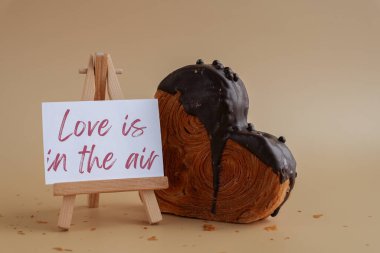 LOVE IS IN THE AIR message on greeting card Delicious fresh croissant topped with Dark chocolate flavor Ganache. Creation unique heart shape bakery. Puff pastry holiday love gift present idea