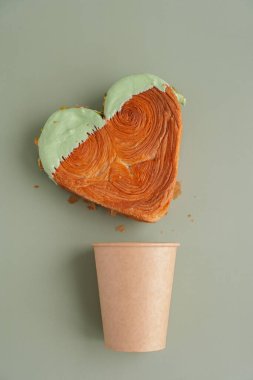Recycled paper coffee cup to go take away with Heart shaped pistachio croissant on green background with copy space. Puff pastry new trendy sweet dessert bakery. Creamy dough experiment 