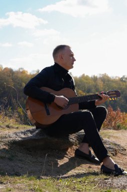 Gülümseyen beyaz ressamın portresi sonbahar parkında ahşap akustik gitar çalar. Genç söz yazarı, sonbahar zamanı dışarıda telli müzik aleti hobisi yapıyor. Sesli müzik doğal olarak iyileşir.