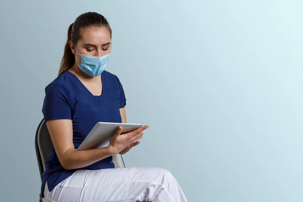 Latina woman, wearing a nurse\'s uniform with mask and mouthpiece, holding a digital tablet in her hands, and a blue background for