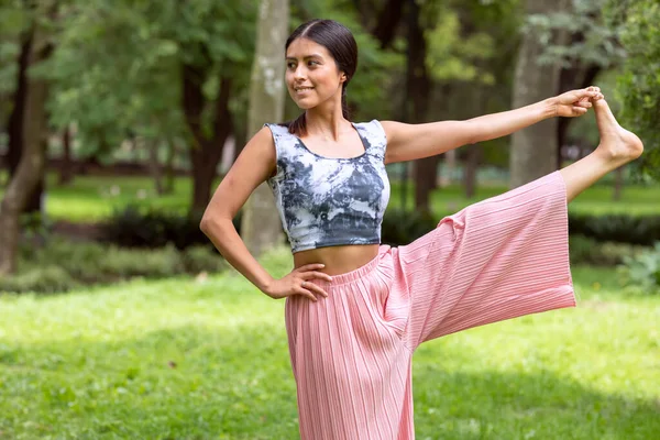 Latin woman doing yoga poses grasping the foot in the park on green grass with a pink outfit