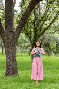 Latin bir kadın parkta bir ağacın kenarında meditasyon yapıyor. Pembe elbiseli yeşil çimlerin üzerinde.