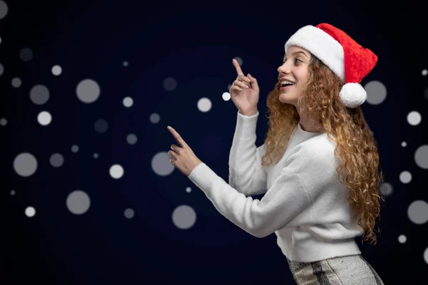 Latino-Frau mit Weihnachtsmütze und Kleidung, die auf einen Raum für Text hinweist, mit unscharfen Lichtern im Hintergrund — Stockfoto