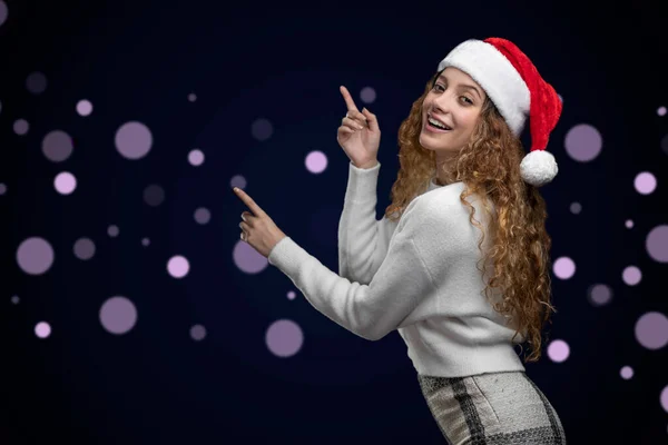 Latino-Frau mit Weihnachtsmütze und Kleidung, die auf einen Raum für Text hinweist, mit unscharfen Lichtern im Hintergrund — Stockfoto