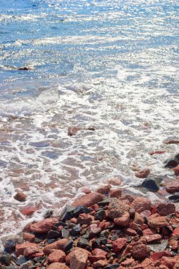 Kumlu bir sahil boyunca yayılmış canlı çakıl taşları, üzerlerinde hafif dalgalar, sakin bir sahil atmosferi yaratıyor.