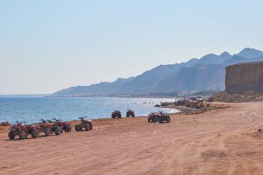 ATV araçları kumlu plaja park edilmiş, arka planda berrak mavi su ve dağlar, sakin bir açık hava macera sahnesi yaratıyor.