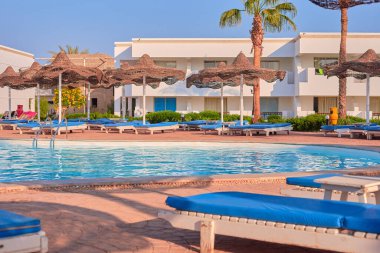 Sakin yüzme havuzu alanında dinlenme koltukları, palmiye ağaçları ve şemsiyeler bulunmakta ve dinlenme ve keyif için rahatlatıcı bir atmosfer yaratmaktadır.