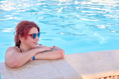 Kadın, sakin su ve parlak güneş ışığıyla çevrili, kollarını kenarda dinlendirip, havuz kenarında boş vakitlerin tadını çıkarıyor.