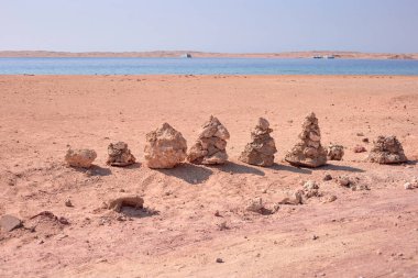 Yığılmış taşlar kumlu sahilde doğal bir heykel oluşturur. Arka planda sakin sular ve sakin atmosferi güçlendiren uzak tekneler vardır.