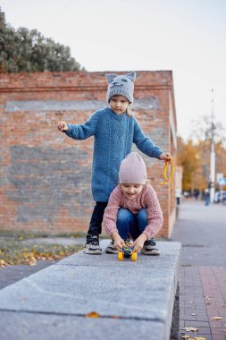 İki genç kız dışarıda oyuncak arabalarla oynuyorlar, sonbahar yaprakları ve kırsal tuğla duvarlarla çevrili, neşeli çocukluk anları sergiliyorlar.