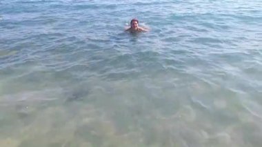 Bir kadın berrak suda enerjik bir şekilde yüzer, su sıçratır ve görünür dalgalar ve güneş ışığı yansımaları ile ferahlatıcı deneyimin tadını çıkarır.