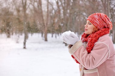 Kışın giyinmiş genç bir kadın, karlı bir manzarada ağaçlarla çevrili, kırmızı bir şapka ve eşarp takarak dışarıda karla oynuyor.