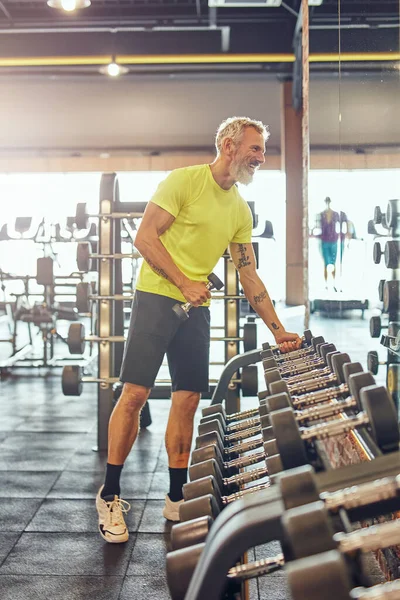 Tiro de comprimento total de um homem atlético maduro feliz em sportswear tomando um halteres, olhando em um grande espelho e sorrindo enquanto se exercita com pesos no ginásio — Fotografia de Stock