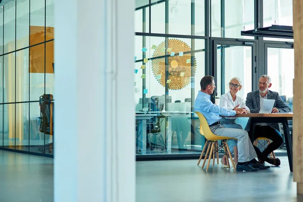 Trois collègues matures assis dans un bureau moderne ou un espace de coworking, utilisant un ordinateur portable et discutant de stratégie d'entreprise — Photo
