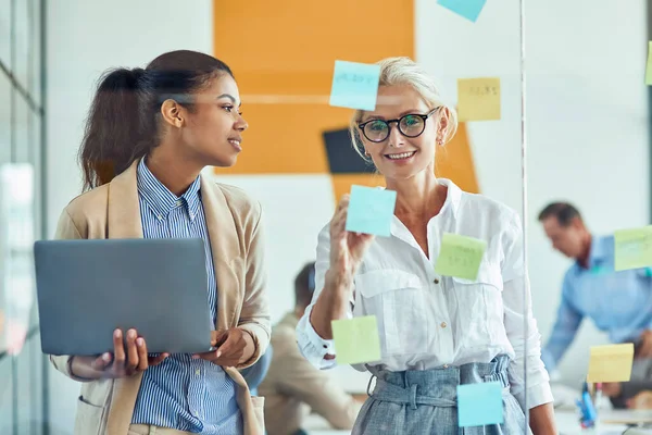 Wo joyeux collègues femmes mettant des notes collantes colorées sur un mur de verre et souriant tout en se tenant dans le bureau moderne, les gens d'affaires travaillant ensemble — Photo