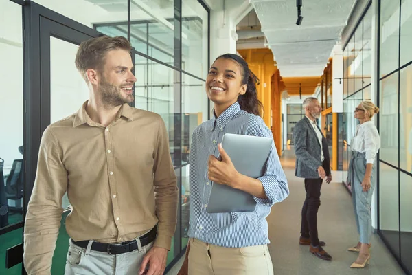 Dos jóvenes felices compañeros de trabajo multirraciales masculinos y femeninos hablando y sonriendo mientras caminan en la oficina moderna — Foto de Stock