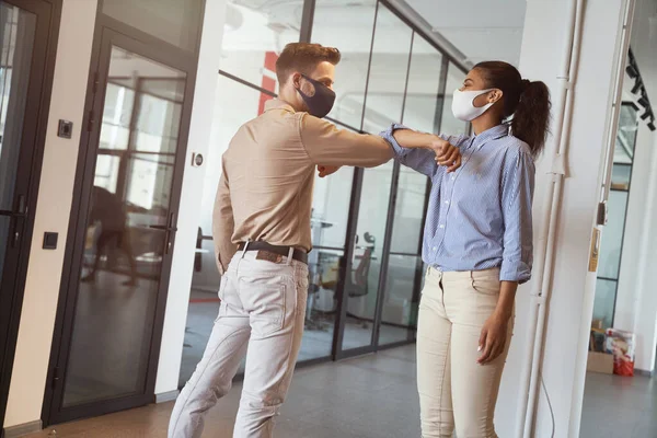 Dos jóvenes y diversos colegas con máscaras protectoras de la cara golpeando los codos, saludándose mientras están de pie en la oficina moderna — Foto de Stock