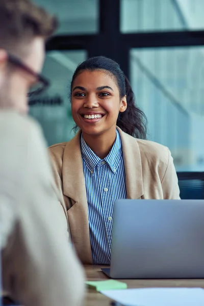 Foto vertikal dari seorang pekerja perempuan ras campuran muda yang ceria yang bekerja pada laptop di kantor modern, berbicara dengan kolega dan tersenyum — Stok Foto