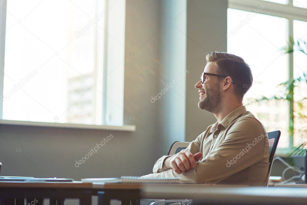 Young happy male office worker sitting at desk in the office and looking aside while having a meeting with colleagues