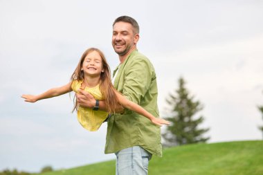 Küçük kızını elinde tutan mutlu bir baba, birlikte eğleniyor. Neşeli kız yeşil parkta babasıyla oynuyor, ellerini uzatıyor, uçuyormuş gibi yapıyor.