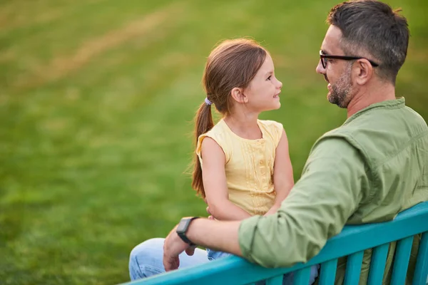 Le jeune père passe du temps avec sa jolie petite fille. Ils se regardent pendant qu'ils sont assis sur le banc dans le parc un jour d'été — Photo