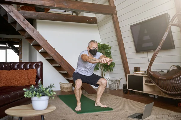 Staying active in self isolation. Middle aged athletic man wearing face protective mask exercising at home while watching online workout on laptop