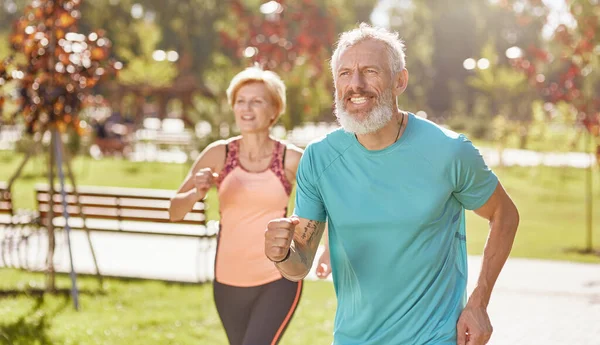 Vorwärts. Aktives, reifes Familienpaar in Sportkleidung, das konzentriert wirkt, während es an einem warmen, sonnigen Tag gemeinsam im Park läuft. Fröhliches Seniorenpaar beim Outdoor-Training — Stockfoto