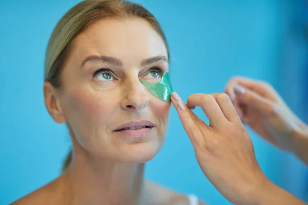 Beauty procedures. Beautiful mature lady looking upside while female beautician putting patches under her eyes