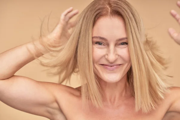 Je me sens joyeuse. Portrait de heureuse femme mature attrayante jouant avec ses cheveux et souriant à la caméra tout en se tenant sur fond beige — Photo