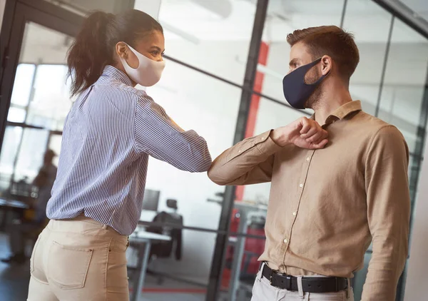 Dos jóvenes colegas de negocios diversos que usan máscaras protectoras de la cara golpeando codos, saludándose mientras trabajan durante la cuarentena covid 19 — Foto de Stock