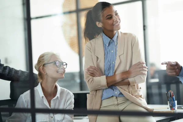 Twee lachende diverse vrouwelijke collega 's kijken opzij terwijl ze een ontmoeting hebben met collega' s in het moderne kantoor — Stockfoto