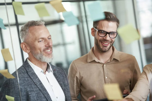 Deux collègues masculins joyeux debout dans le bureau moderne et regardant des notes collantes colorées sur un mur de verre, partageant des idées fraîches — Photo