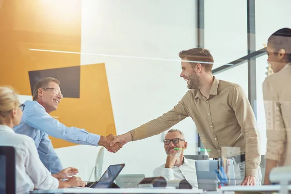 Twee opgewonden zakenmannen schudden elkaar de hand en glimlachen terwijl ze een ontmoeting hebben met collega 's in het moderne kantoor — Stockfoto