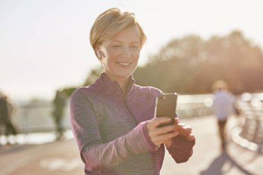 Sonuçlar ortada. Spor giyimli neşeli olgun bir kadının portresi akıllı telefonunu kullanarak güneşli bir günde açık havada koştuktan sonra sonuçları kontrol ediyor.