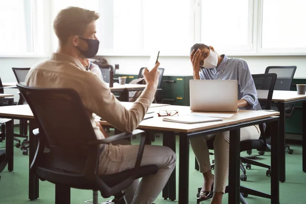 Joven mujer de raza mixta frustrada que usa mascarilla protectora tocando la cabeza mientras está sentada en el escritorio con un colega masculino, trabajando juntos en la oficina durante el brote de covid 19 — Foto de Stock