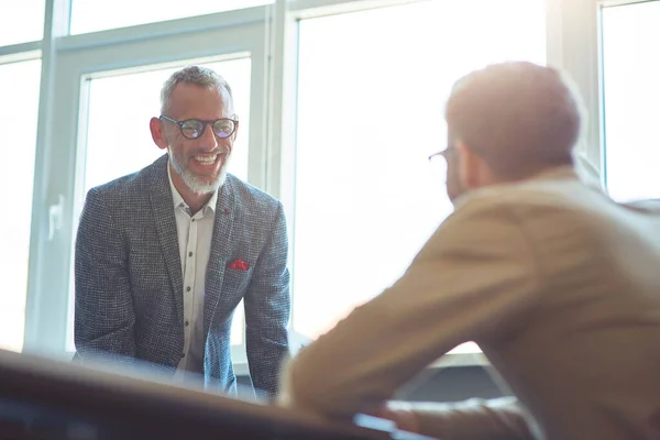 Dojrzały wesoły biznesmen omawia coś ze swoim młodym kolegą i uśmiecha się, pracując razem w nowoczesnym biurze — Zdjęcie stockowe