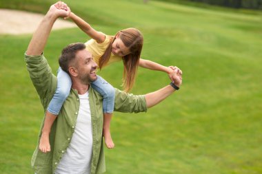 Mutlu bir baba, yeşil alanda dururken küçük kızıyla birlikte onu omuzlarında tutarak eğleniyor, kızı ellerini uzatıyor, uçuyormuş gibi yapıyor.