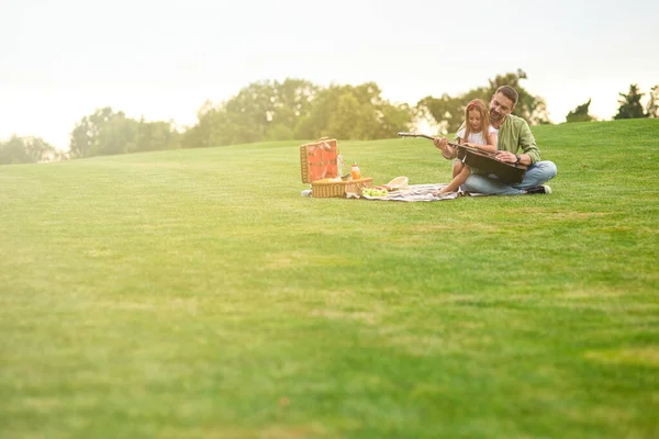 Plan complet de la petite fille heureuse assise avec son père aimant sur une herbe verte dans le parc et apprenant à jouer de la guitare — Photo