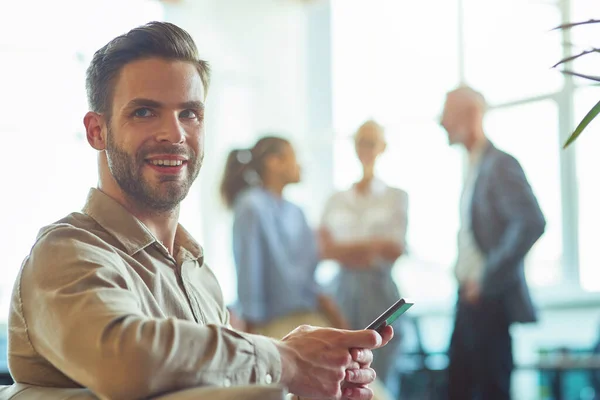 Young happy businessman sitting in the modern office, holding smartphone and smiling at camera while his colleagues talking on the background