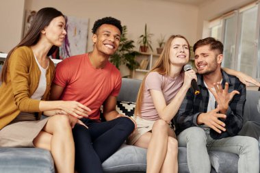 Karaokeyi tecrübe et. Evde karaoke yapan, mikrofonla şarkı söyleyen, oturma odasındaki kanepede otururken birlikte harika vakit geçiren mutlu genç çok kültürlü arkadaşlar.