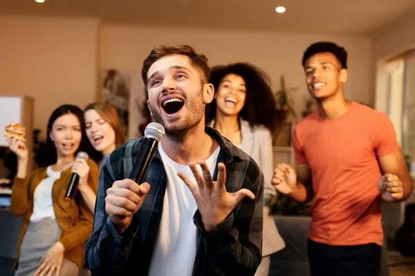 Evde eğlenmek. Hevesli yakışıklı genç adam mikrofonla şarkı söylerken içeride arkadaşlarıyla karaoke yapıyor, modern dairede duruyor.