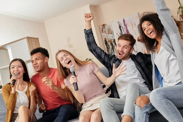 Feel the music. Group of multicultured friends looking excited while singing with microphone, playing karaoke at home, sitting on the couch in the modern apartment