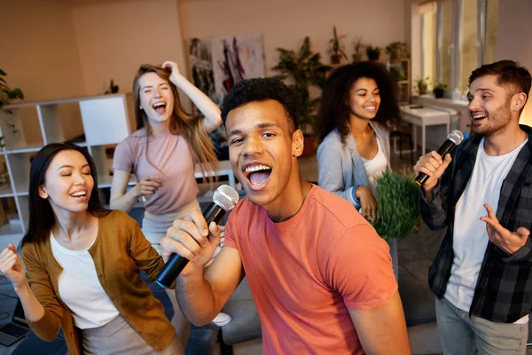 Radiasi kesenangan. Pemuda yang antusias bernyanyi dengan mikrofon saat bermain karaoke dengan teman-teman di apartemen modern, mengadakan pesta di rumah — Stok Foto