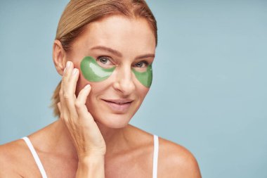 Mavi stüdyo duvarında hidrojel göz bantları olan mutlu beyaz kadın.