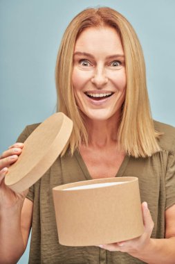 Mutlu olgun bir kadının dikey portresi hediye kutusunu açıyor ve şaşırmış bir yüz ifadesiyle kameraya bakıyor.