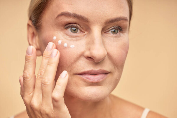 Close up of beautiful mature woman applying anti-aging cream under the eyes, isolated on beige background