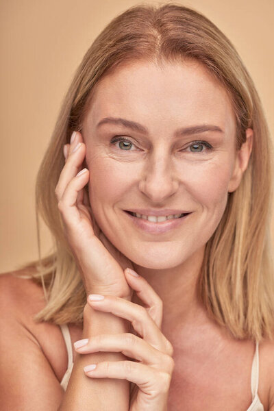 Vertical shot of a beautiful middle aged caucasian woman touching her clean and soft skin, looking at camera while posing over beige background in studio